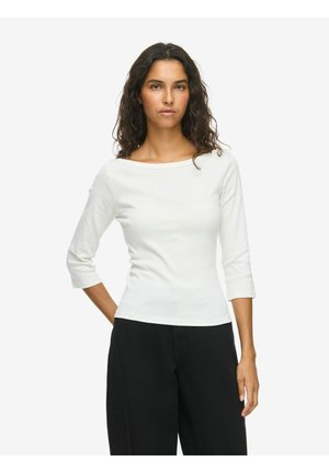 Femme aux longs cheveux bouclés et foncés portant un haut blanc à manches trois-quarts et un pantalon noir, debout avec une main derrière le dos.
