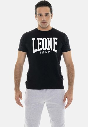 Mann steht frontal, trägt ein schwarzes "LEONE 1947" T-Shirt und hellgraue Jogginghosen vor weißem Hintergrund.