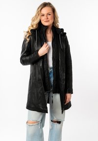 Zwarte leren jas met een ritssluiting aan de voorkant, verlengd silhouet, hoge kraag en textuurdetails, gedragen over een witte top en lichtblauwe, gescheurde jeans.