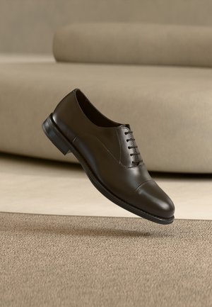 Brun lærsko med glatt tekstur, snøring, avrundet tå og en lav svart gummihæl, vist mot en nøytral bakgrunn.