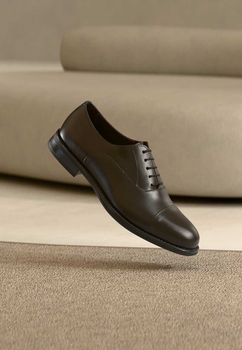 Καφέ δερμάτινο παπούτσι με λεία υφή, σχέδιο με κορδόνια, στρογγυλεμένη μύτη και χαμηλό μαύρο λαστιχένιο τακούνι, τοποθετημένο σε ουδέτερο φόντο.