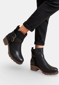 Bottes noires à la cheville avec de grosses semelles brunes et des sangles décoratives à boucles, portées avec un pantalon noir retroussé.