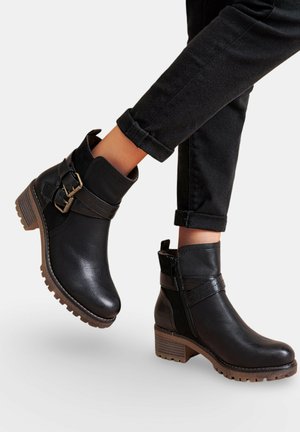 Bottes noires à la cheville avec de grosses semelles brunes et des sangles décoratives à boucles, portées avec un pantalon noir retroussé.