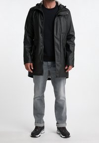 A man stands wearing a black waterproof jacket, dark sweater, gray jeans, and black sneakers, against a plain white background.