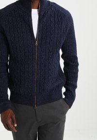 Marinblå kabelstickad cardigan med full dragkedja, ribbade muddar och nederkant, tillverkad av en texturerad ullblandning. Har en hög krage för extra värme.