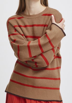 Kobieta w beżowym dzianinowym swetrze z czerwonymi paskami, skrzyżowanymi ramionami, trzymająca lewą górną część ramienia na jednolitym tle.