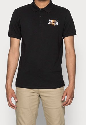 Model wearing a black polo shirt with a colorful "Jack & Jones" logo, paired with beige pants, standing against a plain background.