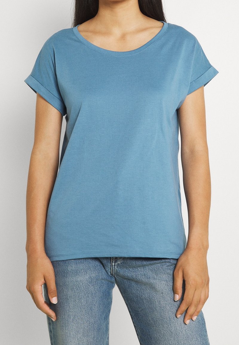 Femme portant un t-shirt bleu clair à manches courtes avec des revers roulés et un jean bleu, debout devant un fond blanc uni.