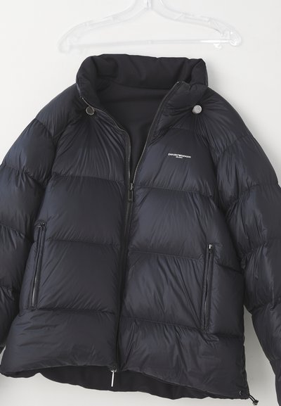 Black puffer jacket with a high collar, horizontal quilted design, zippered front and pocket, featuring a logo on the left chest.
