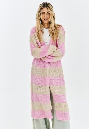 Uśmiechnięta kobieta z długimi blond włosami, ubrana w długi, gruby sweter kardigan w szerokie różowe i beżowe paski, biały top i luźne spodnie.