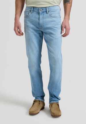 Mann iført lyseblå straight-fit jeans og brune snøresko, stående mot en ensfarget lys bakgrunn.