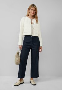 Biała kurtka typu cropped z teksturowanej tkaniny, z przednimi kieszeniami i złotymi guzikami, zestawiona z ciemnoniebieskimi spodniami typu wide-leg i beżowymi loafersami.