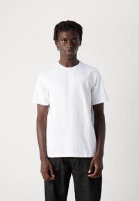 Jonge man met korte dreadlocks, gekleed in een eenvoudig wit T-shirt en zwarte jeans, staand tegen een effen lichte achtergrond.