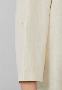 Beige Leinenhemd mit umgeschlagenem Ärmel, glatter Textur und einem kleinen Knopfdetail nahe dem Bund. Einfaches und elegantes Design.