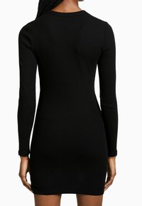 Svart ribbad långärmad klänning med en figursydd design, strukturerad tyg och rundad halsringning; fållen slutar vid mitten av låret.