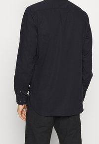 Černá košile s dlouhými rukávy z hladké látky, s knoflíkovým manžetem a zadní vsadkou. Mírně zakulacený spodní lem a minimalistický design.