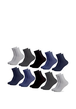 Person zieht schwarzen, gerippten Socken auf nackten Fuß an, trägt hochgekrempelte dunkle Hose und gestreiftes Langarmshirt mit Perlenarmband.