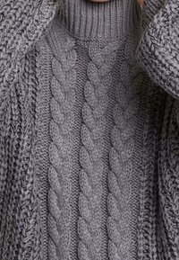 Pull en tricot épais gris avec des motifs en tresses et un col roulé côtelé, partiellement recouvert par des manches assorties en tricot épais.