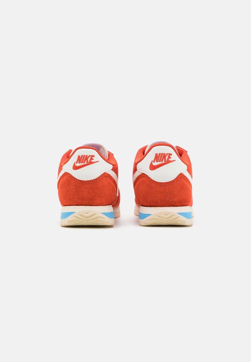 Zapatillas Nike de ante rojo y malla con acentos en blanco, que presentan una lengüeta en azul y suela de goma texturizada, vistas desde la parte trasera.