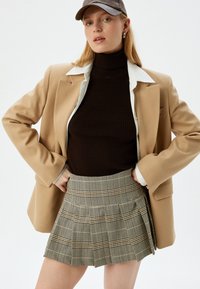 Beige blazer, aansluitende bruine coltrui en een geplooide geruite rok met bruine en gele lijnen. Een pet maakt de outfit compleet.