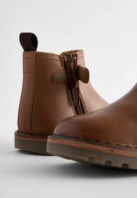 Bruna läderankelboots har en sidoglid, texturerad tygfoder, en dragflik och en hållbar gummisula med synliga sömmar.