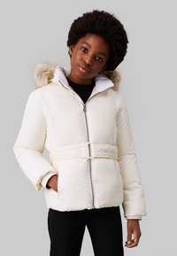 Giacca puffa bianca con cappuccio in pelliccia sintetica, vita con cintura e chiusura a zip. Progettata con maniche lunghe e accenti neri sul colletto.