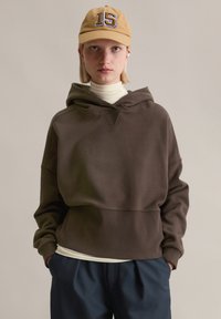 Braunes Kapuzen-Sweatshirt mit lockerem Schnitt, strukturierter Stoff und V-förmigem Ausschnitt, getragen über einem hellfarbenen Rollkragenpullover und einer dunkelblauen Hose.