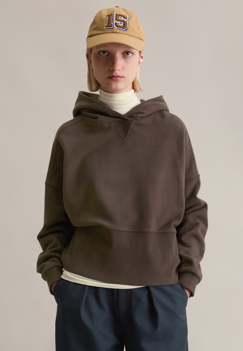 Braunes Kapuzen-Sweatshirt mit lockerem Schnitt, strukturierter Stoff und V-förmigem Ausschnitt, getragen über einem hellfarbenen Rollkragenpullover und einer dunkelblauen Hose.