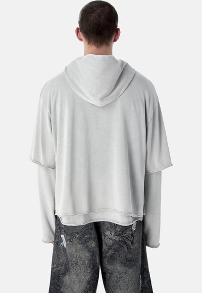 Homme de dos portant un sweat à capuche gris clair superposé et un jean noir délavé et usé, sur fond blanc.
