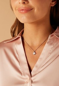 Zilveren bloemenhanger ketting op een delicaate ketting, met een gestructureerd bloemdesign, gedragen over een glanzende roze satijnen blouse.