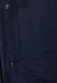 Veste rembourrée bleu marine avec une texture lisse, fermeture éclair et accents de boutons ; détail de couture visible le long des coutures.