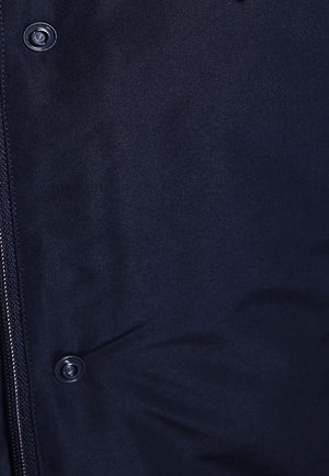 Veste rembourrée bleu marine avec une texture lisse, fermeture éclair et accents de boutons ; détail de couture visible le long des coutures.