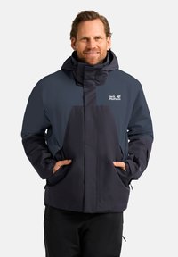 Giacca impermeabile da uomo in blu navy scuro e nero, dotata di tasche con zip, polsini regolabili e colletto alto con cappuccio.