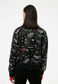 Giubbotto bomber nero con testo grafico bianco e rosso e scarabocchi, polsini e vita a costine, dal tessuto liscio e lucido.