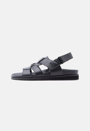 Sandalia de cuero negro con suela plana, correas ajustables sobre el pie y alrededor del talón, y una zona delantera cerrada.