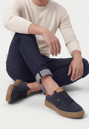 Homme assis en tailleur portant un jean bleu foncé retroussé, une chemise beige à manches longues et des baskets bleu marine avec des semelles et des accents marron.