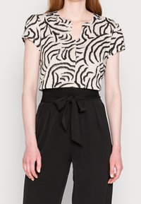 Haut à manches courtes en tissu beige à rayures zèbre noires, avec un col en V et une coupe décontractée, associé à un pantalon noir taille haute.