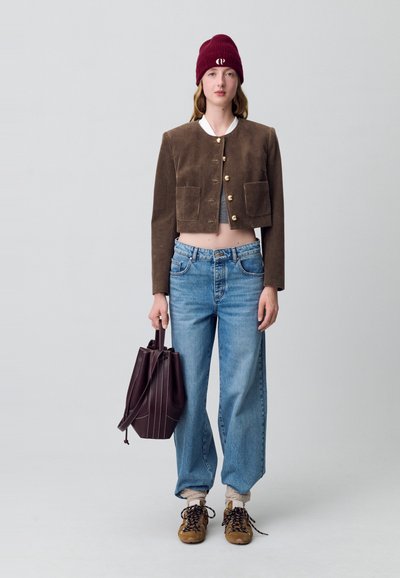 Giacca cropped in corduroy marrone con bottoni dorati, maglione grigio a maglia, jeans blu a tutto tondo, sneakers marroni e un berretto bordeaux. Tiene una borsa scura.