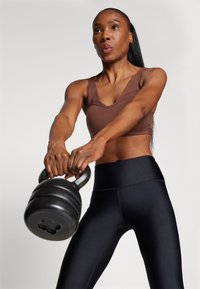 Smeđa sportska grudnjak i crne tajice visokog struka koje nosi osoba koja drži crnu kettlebell. Glatke teksture i prilagođen dizajn.