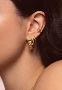 Orecchini a cerchio in oro con pietre di vetro colorate—verdi, rosa e multicolore—mostrano intricati motivi e texture. Sono visibili i fori nelle orecchie.