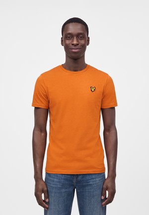 Homme portant un t-shirt orange à manches courtes avec un petit logo d'oiseau jaune sur la poitrine, associé à un jean bleu, debout devant un fond uni.