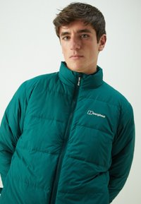Teal isoleret jakke med lynlås og høj krave. Har quiltet mønster og broderet logo på brystet.