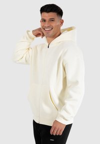 Krämsfärgad zip-up hoodie i mjukt tyg, med en känguruficka, ribbade muddar och en rymlig huva. Modellen står mot en grå bakgrund.