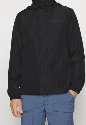 Mężczyzna w czarnej kurtce z kapturem i elastycznymi mankietami oraz niebieskich spodniach bojówkach, stojący z rękami swobodnie opuszczonymi wzdłuż ciała.