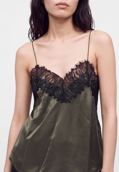 Femme portant un haut caraco en satin vert olive avec fines bretelles et dentelle noire à l'encolure, sur fond blanc uni.