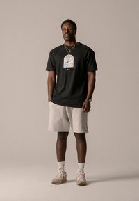 Svart bomulls-T-skjorte med grafisk trykk, kombinert med lysegrå shorts og beige joggesko. Tilbehør inkluderer halskjede og armbåndsur.