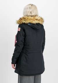 Veste noire isolée avec une capuche doublée de fourrure. Comprend un patch du drapeau américain, des poches latérales et une étiquette en tissu rouge sur le bras.