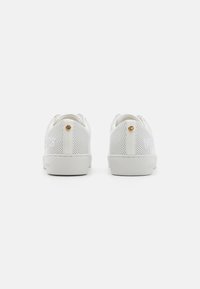 Hvide perforerede sneakers med en glat gummisål, der har et gyldent logoaccent på hælen og subtil branding på siden.