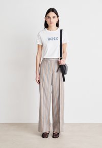 Balta marškinėliai su „BOSS“ mėlynos spalvos užrašu, derinami su šviesiai beige ir šviesiai mėlynomis raištinėmis plačiomis kelnėmis. Modelis laiko mažą juodą rankinę.