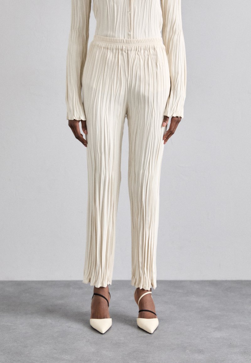 Pleated ivory trousers with an elastic waistband, featuring a relaxed fit. Paired with pointed-toe heels with a black ankle strap.